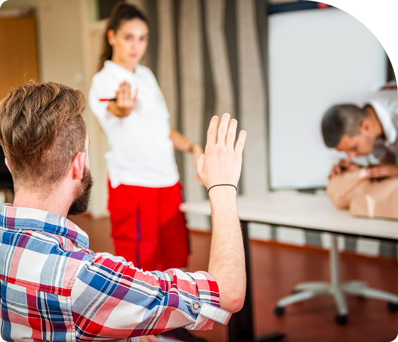 A person in a plaid shirt raises a hand during a training session, while an instructor gestures with a pen, and another individual practices CPR on a mannequin in the background.